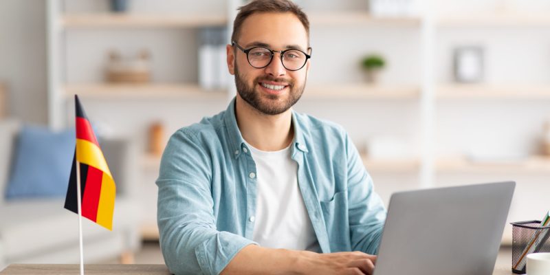 Happy young guy sitting at table with flag of Germany, using laptop pc, studying foreign language online at home. Modern remote education, abroad work and citizenship concept